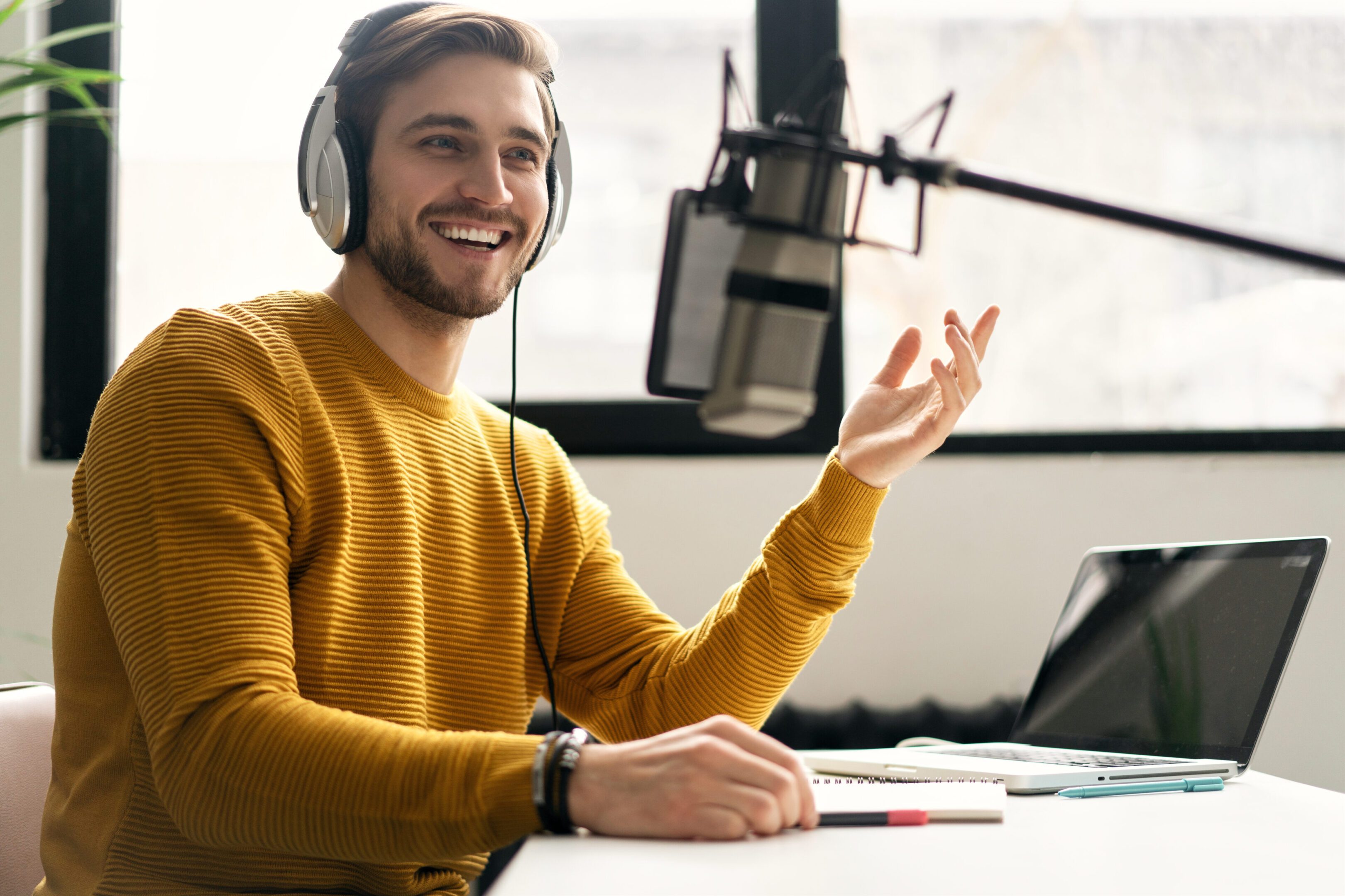 Person in a yellow sweater sitting at a desk with a microphone, headphones, and a laptop, gesturing while speaking.